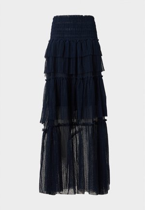 Femme aux longs cheveux raides portant un top polo en maille noire à manches courtes et une jupe maxi en tulle noir superposée, les mains dans le dos.
