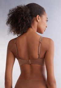 Hnědá podprsenka s nastavitelnými ramínky a zapínáním na háček, vyrobená z hladkého materiálu. Má bezešvý design s minimalistickým vzhledem.