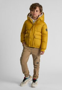 Doudoune jaune avec capuche, deux poches avant et un patch logo. Pantalons cargo beiges avec poches latérales et chaussures de sport noires.