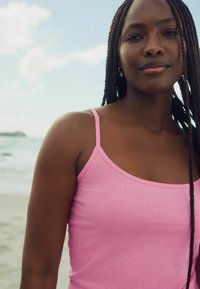 Top de algodón rosa sin mangas con tirantes delgados, textura suave y corte ajustado. Cabello trenzado y fondo oceánico visibles.