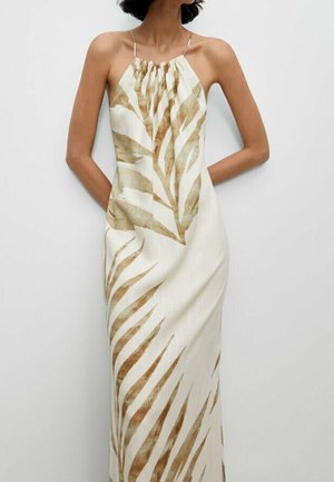 Woman wearing a sleeveless, cream maxi dress with thin straps and large brown tropical leaf prints, standing with hands behind back.
