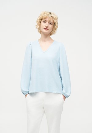 Žena s krátkymi kučeravými blond vlasmi, oblečená v svetlomodrej blúzke s výstrihom do V a dlhými rukávmi, biele nohavice, ruky vreckách, neutrálne pozadie.