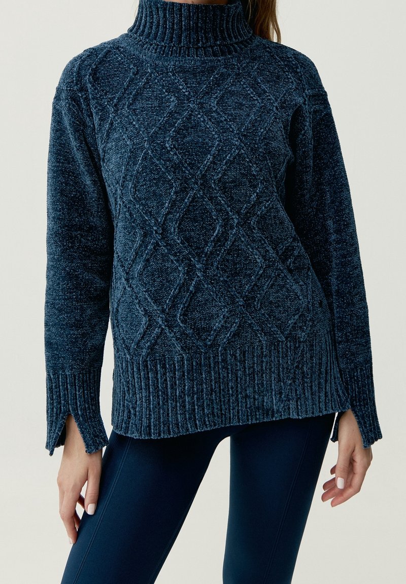 Mörkblå stickad tröja med hög krage, texturerat diamantmönster och ribbad fåll. Sidossprund ökar komfort och passform.