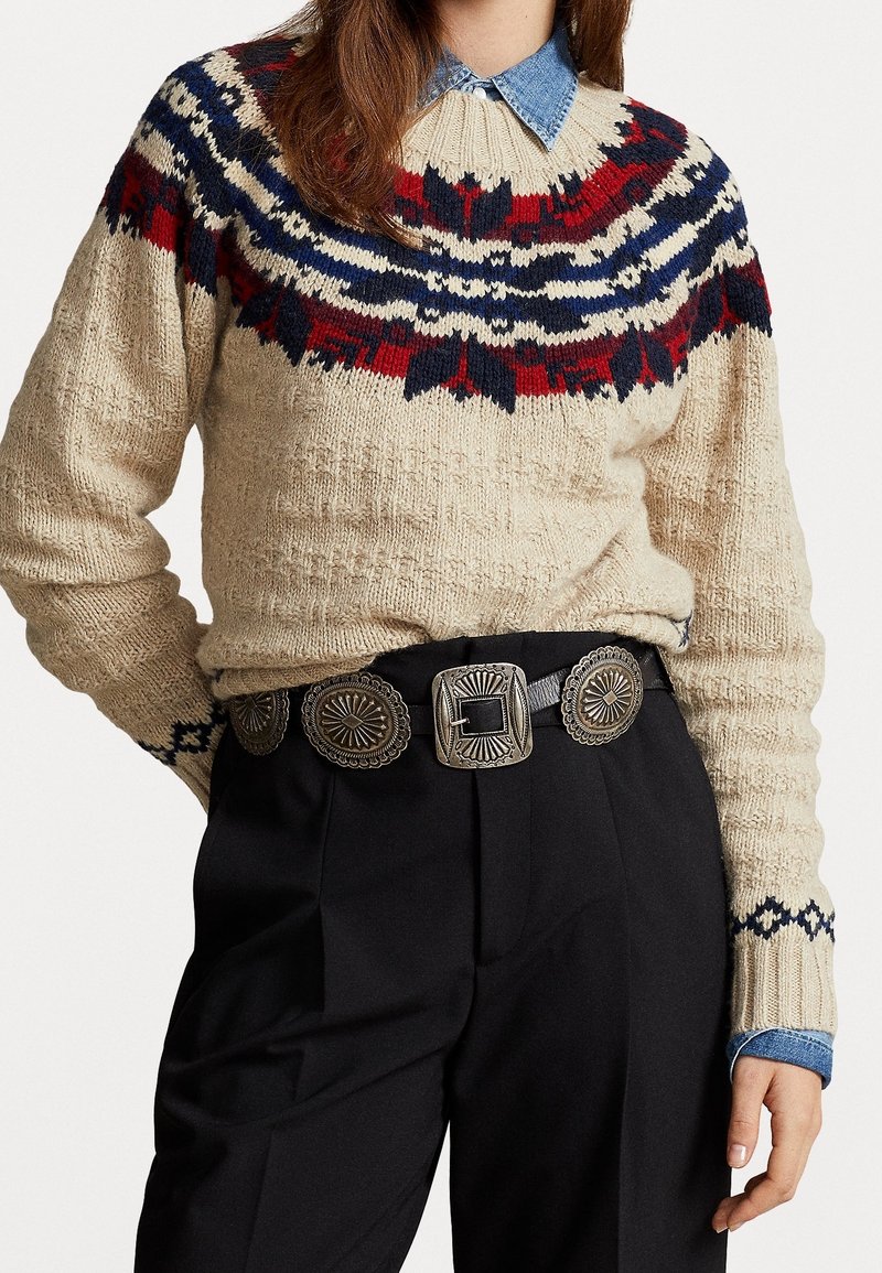 Suéter de punto beige con un cuello nórdico de patrón colorido, combinado con pantalones negros y un cinturón decorativo plateado con acentos circulares.