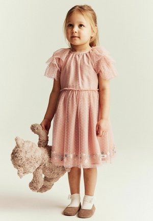 Junges Mädchen im rosa Kleid mit Rüschen, das einen großen Plüsch-Teddybären hält und auf hellem Hintergrund mit weißen Socken und braunen Schuhen steht.