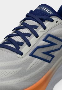 Κοντινό πλάνο ενός αθλητικού παπουτσιού σε μπεζ και μπλε χρώμα, με μπλε κορδόνια, υφή πλέγματος στο πάνω μέρος και πορτοκαλί ενδιάμεση σόλα.