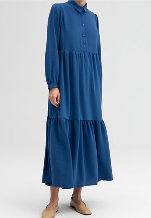 Femme portant une longue robe bleue à boutons et à étages avec des manches longues, ainsi que des chaussures beiges à talons bas, debout devant un fond uni.