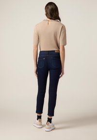 Top beige a maniche corte con design a chiave sul retro, abbinato a jeans scuri su misura con cuciture decorative e tasche.