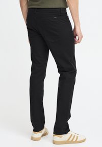 Pantalons en denim noir à coupe slim, dotés de deux poches arrière et d'un logo sur la taille, associés à des baskets blanches et beige.