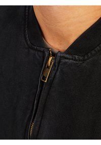 Czarna kurtka bombowa z złotym zamkiem błyskawicznym, prążkowanym kołnierzem i teksturowanym materiałem. Dolna część kołnierza jest widoczna nad beżową koszulką.