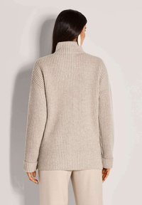 Frau mit langen dunklen Haaren, die einen beigen gerippten Rollkragenpullover und passende Hosen trägt, mit dem Rücken zu einem schlichten, hellen Hintergrund gewandt.