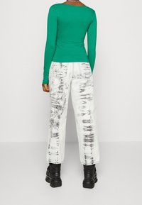 Grünes langärmliges enges Oberteil, kombiniert mit weißen Jeans mit einem schwarzen Batikmuster, getragen mit schwarzen klobigen Stiefeln und einer schlichten Halskette.