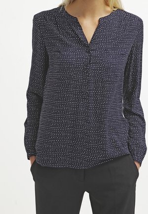 Femme portant un chemisier bleu marine à manches longues avec un petit motif à pois blanc et un pantalon noir, mains dans les poches, debout devant un fond blanc.