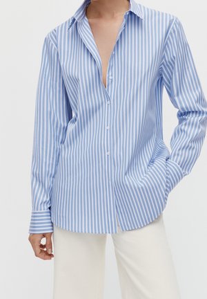 Person wearing a light blue and white vertical striped button-up shirt with long sleeves and beige pants, hand in pocket, against white background.