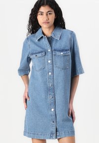 Δημιουργία από τζιν σε ανοιχτό μπλε χρώμα με κοντά μανίκια, κουμπί κλεισίματος, δύο μπροστινές τσέπες και ευθεία σιλουέτα. Λείος υφάσματος και κλασικός σχεδιασμός.