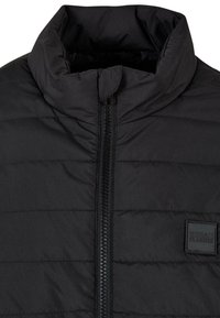 Black padded jacket with a high collar, featuring horizontal quilted lines, a full zipper, and a small branded patch on the left side.