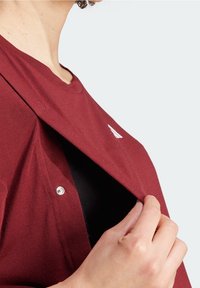 Camisa burdeos con cierre a presión, con una capa subyacente negra. Un logo blanco es visible en la tela. Material de algodón texturizado.