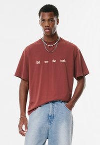 T-shirt couleur rouille avec coupe oversize, arborant le texte "Dis-moi la vérité" en crème. Porté avec un jean en denim bleu clair et des colliers superposés.