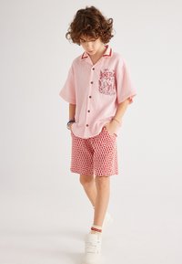 Kortebloesem shirt in het roze met rode accenten en een grafische voorzak, gecombineerd met shorts met rode patronen. Witte sneakers maken de outfit compleet.