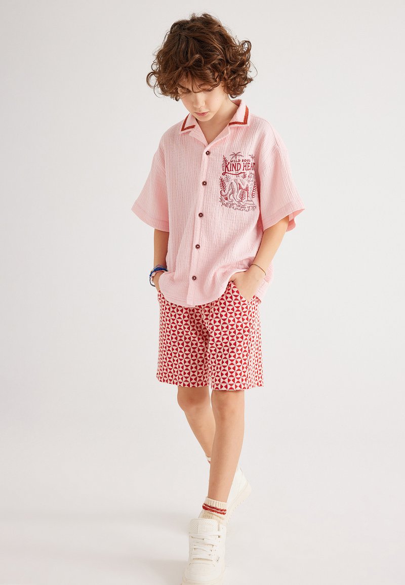 Kortebloesem shirt in het roze met rode accenten en een grafische voorzak, gecombineerd met shorts met rode patronen. Witte sneakers maken de outfit compleet.