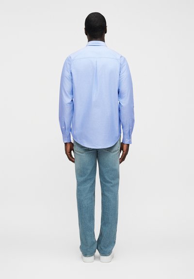 Ανοιχτό μπλε πουκάμισο με μακριά μανίκια, εφαρμοστή γραμμή, ίσια τελείωμα και μανσέτες με κουμπιά, σε συνδυασμό με ανοιχτόχρωμο τζιν και λευκά αθλητικά παπούτσια.