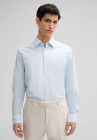 Hellblaues Langarm-Hemd mit Knöpfen, weiche Textur, spitzer Kragen, weiße Knöpfe, kombiniert mit beigen Anzughosen. Einfaches, klares Design.