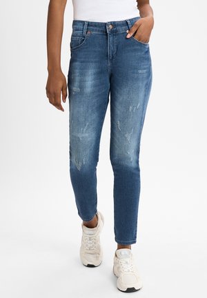 Blue denim jeans with a fitted design, featuring distressed details and a five-pocket style. Worn with white sneakers.