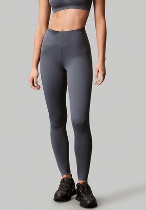 Femme portant un legging gris foncé taille haute, un soutien-gorge de sport assorti et des baskets noires, debout devant un fond gris uni.