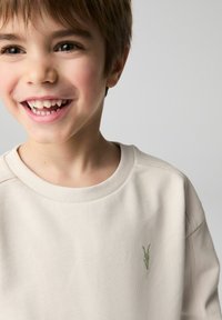 Beige sweatshirt med ribbede kanter og et lille grønt broderet logo på brystet, tekstureret stof, båret af et smilende barn.