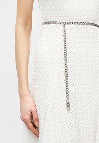 Robe blanche texturée avec une fine ceinture chaîne argentée tombante, arborant un fermoir avec les initiales « MK » près de la taille.