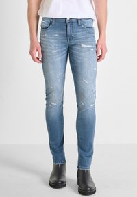 Jeans skinny de denim azul desgastados con detalles rasgados, diseño de cinco bolsillos y cremallera, combinados con botas negras hasta el tobillo.