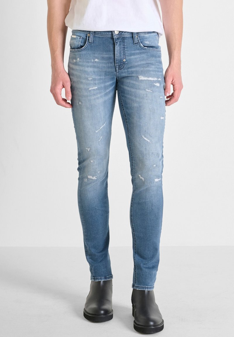 Jeans skinny de denim azul desgastados con detalles rasgados, diseño de cinco bolsillos y cremallera, combinados con botas negras hasta el tobillo.