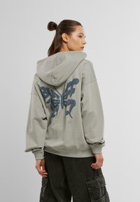 Overdimensioneret grå hoodie med et iøjnefaldende blåt sommerfugleprint på ryggen. Har snoretræk i huen og ribbede mansjetter.