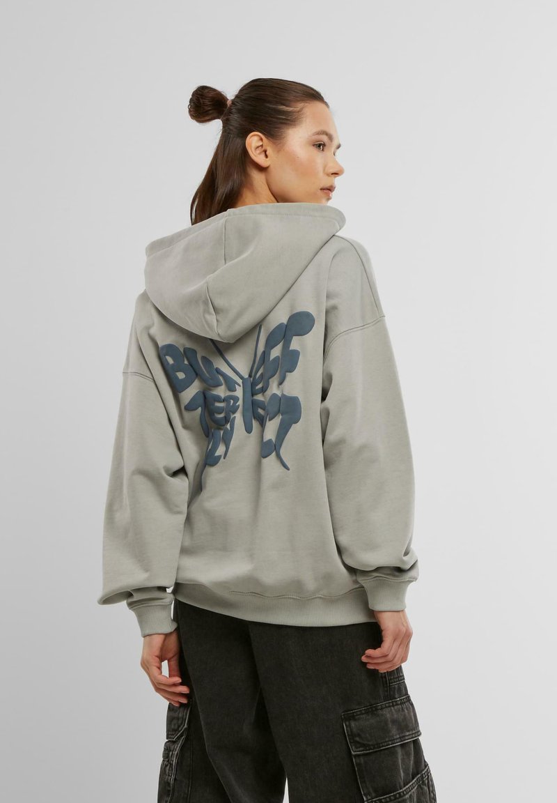 Hoodie gris surdimensionné avec un impressionnant motif de papillon bleu à l'arrière. Comprend une capuche avec cordon de serrage et des poignets côtelés.