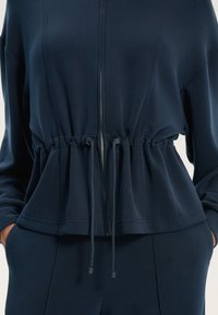 Mornarčko plava majica sa zipom, cinčana u struku, s trakama za podešavanje i glatkom teksturom. Sužene rukave i suptilne detalje šivanja.