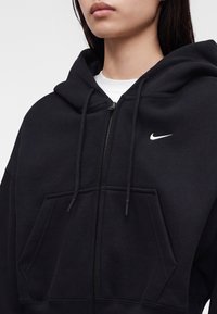 Близък план на човек, носещ черно худи с цип Nike с връзки и бяло лого на гърдите върху бяла риза.