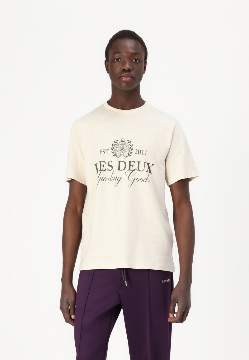 Beige bomull t-skjorte med korte ermer, rund hals, med svart trykk "LES DEUX Sporting Goods" og "EST. 2011" på fronten.