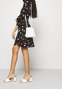 Kobieta w czarnej sukience w kwiaty, nosząca białe czółenka na obcasie oraz trzymająca małą białą torebkę na ramię, na jednolitym jasnym tle.