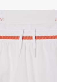 Shorts sportivi bianchi con una vita testurizzata caratterizzata da un accento a strisce rosse e arancioni. La vita presenta chiusure con cordino e dettagli in rete.
