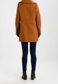 Brauner Wollmischmantel mit hohem Kragen, tailliertem Schnitt und Reißverschlussärmel, kombiniert mit engen marineblauen Jeans und schwarzen Ankle Boots.