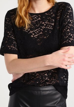 Femme portant un haut à manches courtes en dentelle florale noire avec une sous-couche noire unie, associé à un pantalon en cuir noir.