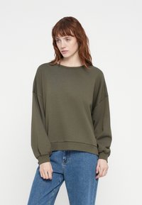 Olive green sweatshirt with long sleeves, round neckline, and ribbed hem. The fabric appears soft and slightly textured. Worn with blue jeans.