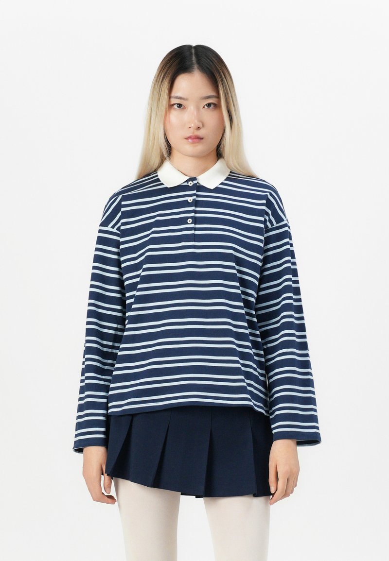 Marine- og lyseblå stribet langærmet polo-shirt med hvid krave og tre-knaps lukning, parret med en marinefarvet plisseret nederdel.