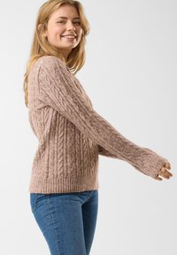 Sweter w warkocze w delikatnym różowym kolorze, z prążkowanym dołem i szerokimi rękawami. Nosi się go z dopasowanymi niebieskimi dżinsami, które ukazują szczegóły faktury materiału.