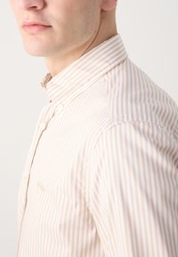 Camicia con colletto caratterizzata da righe verticali beige e bianche, chiusura a bottoni e una morbida trama di cotone. Dettaglio del logo ben visibile sul petto.