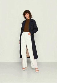 Casaco trench azul-marinho sobre uma blusa castanha, combinado com calças largas brancas e sandálias de plataforma claras; apresenta botões e uma cintura cingia.