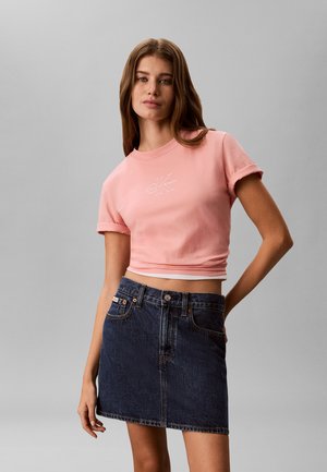 Νεαρή γυναίκα με μακριά καστανά μαλλιά, που φοράει ροζ κοντό T-shirt και σκούρη μπλε τζιν φούστα, στέκεται μπροστά σε απλό γκρι φόντο.