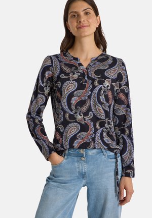 Femme portant un chemisier sombre à manches longues avec un motif paisley coloré et un jean bleu clair, debout avec une main dans la poche.