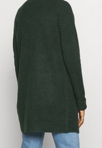 Osoba oblečená v tmavě zeleném dlouhém pleteném kardiganu s ohrnutými manžetami a světle modrých džínách, zobrazená ze zadní strany.