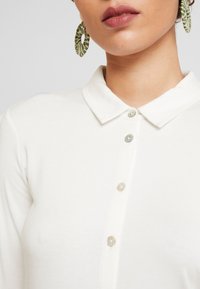 Weißes Poloshirt mit spitzem Kragen, ausgestattet mit hellen Knöpfen. Große grüne, strukturierte Creolen ergänzen den Ausschnitt.
